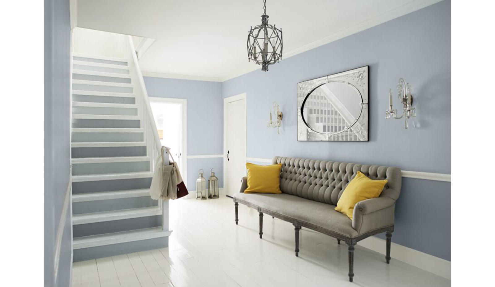 Área de estar azul-gris con escaleras de dos tonos, sofá gris y cojines amarillos, lámpara de araña 