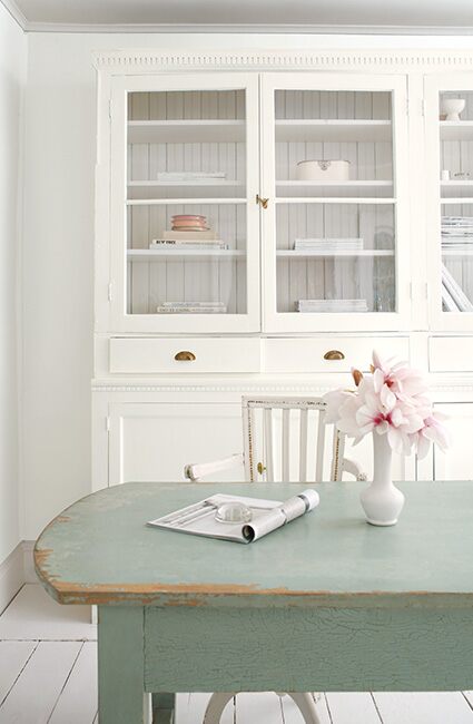 Comedor shabby chic: paredes blancas, armario, silla; mesa azul, flores; piso de madera gris claro.